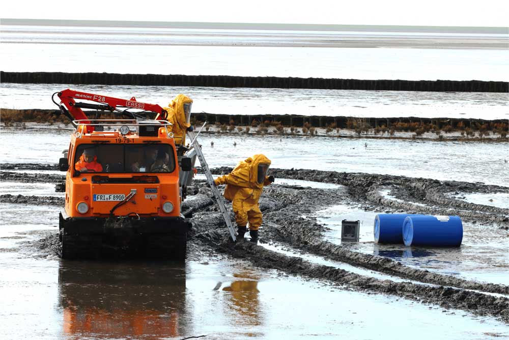 Einsatzkräfte in Chemikalienschutzanzügen üben die Bergung von Fässern mit Gefahrgut aus dem Watt